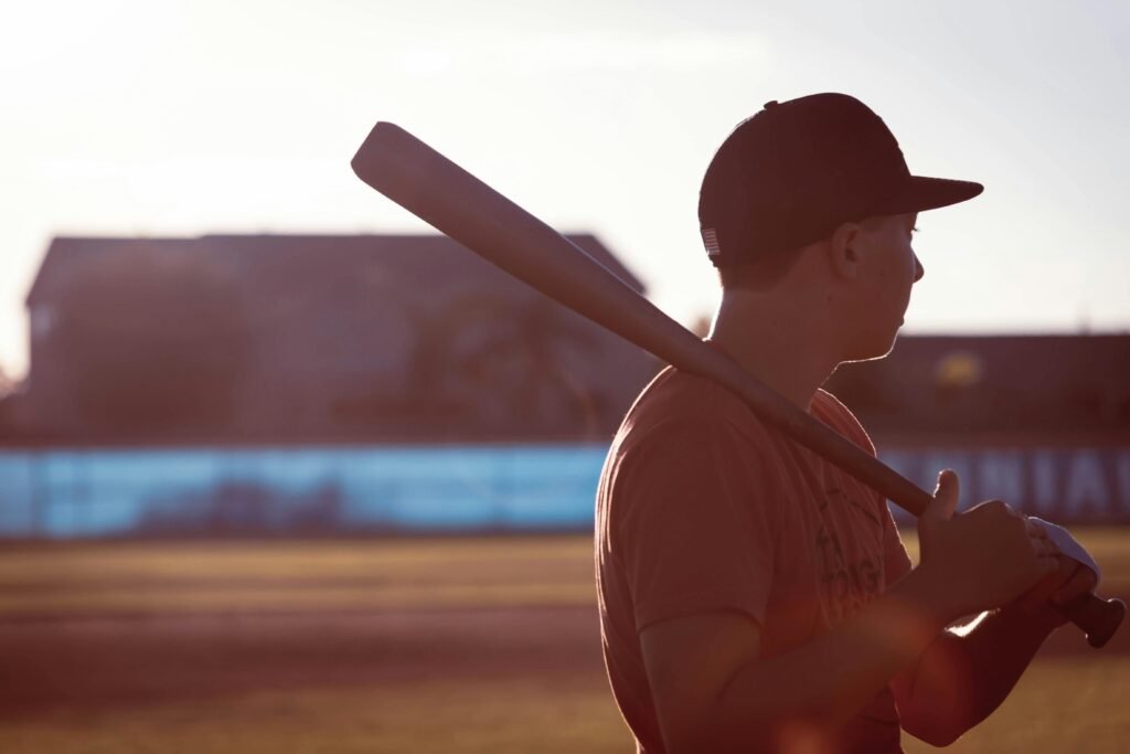 A teenage boy holding a baseball bat looks towards the field at sunset, ready to play.